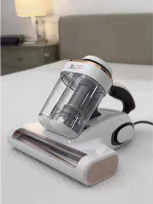 A close-up shot of a white and clear handheld vacuum cleaner on a white bed. The vacuum cleaner has a cylindrical, clear plastic dust container and a brush head. The bed is neatly made with a gray headboard in the background.