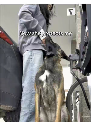 A woman stands at a gas pump with her dog standing up near her. The dog is tall and has markings.