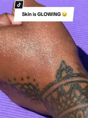Close-up of a hand and part of a wrist, showcasing glowing skin. The skin has a shiny, radiant appearance, indicative of a skincare product application. A detailed, dark tattoo adorns the wrist. The background is a solid, light purple.