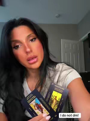 A woman with dark hair and fair skin is holding two tarot cards close to the camera. The cards are partially visible, with one featuring a figure in a robe and the other showing an image of a figure on a road. The woman's facial expression is serious and focused.
