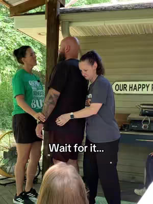 A group of three people are standing outdoors. The focus is on a man in the middle, facing away from the camera, with two women on either side of him, one hugging him. Text overlayed reads 'Wait for it...'