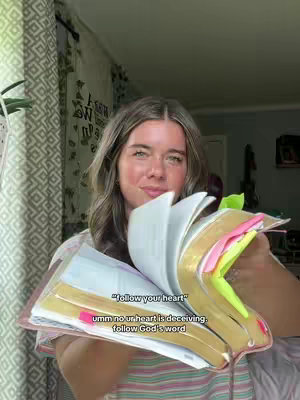 A young woman with long wavy hair, smiling slightly, holding an open Bible with colorful page markers. She stands in front of a background with a patterned curtain and some plants. Overlaying her is text that seems to be a response to the caption.