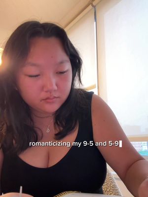 The cover image features a close-up of a person, likely a young woman, sitting indoors. She is looking down with a focused expression. She wears a black tank top and a necklace with a pendant. Natural light enters from the right side of the frame, possibly a window. The background is simple with some blurred objects. Text added to the cover image reads, "romanticizing my 9-5 and 5-9"