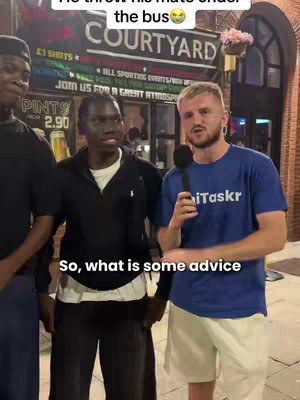 A street interview scene featuring a man wearing a blue t-shirt with "iTaskr" printed on it, asking a question into a microphone. Two other men are in the background, one wearing a black outfit, the other in a dark jacket, both looking toward the interviewer. The background shows a bar or pub with a sign that reads "Courtyard."