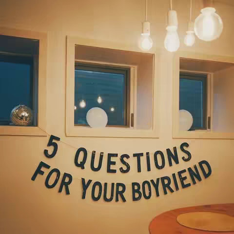 The image shows a warm-toned indoor scene with string lights hanging in front of windows. A banner with the text "5 QUESTIONS FOR YOUR BOYFRIEND" hangs below the windows. A round table is prominent in the foreground.