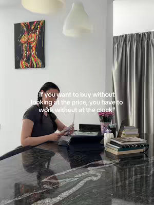 A woman sits at a table with a laptop, books, and other items, holding money, with a painting on the wall and text overlaid.