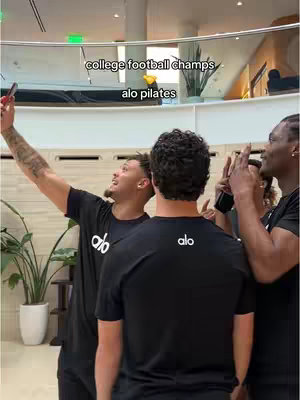 Three men are posing for a photo, with one holding up a phone for a selfie. They are wearing black t-shirts. Text overlays read 'college football champs' and 'alo pilates'.