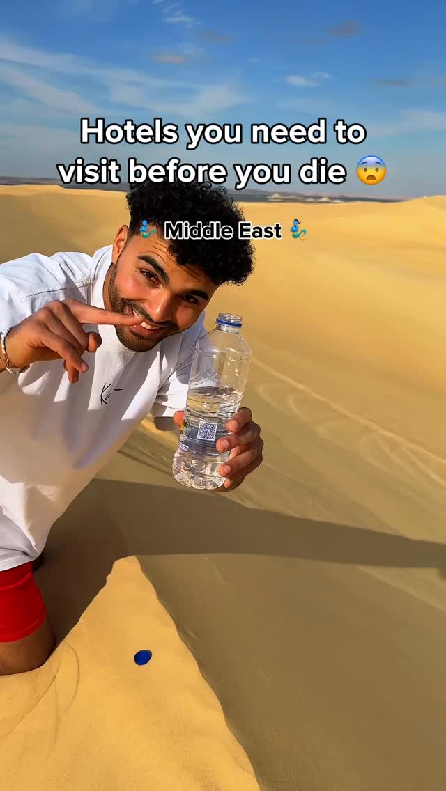 A selfie of a smiling man in a white shirt holding a clear water bottle in a desert environment under a blue sky. The background is a sand dune.
