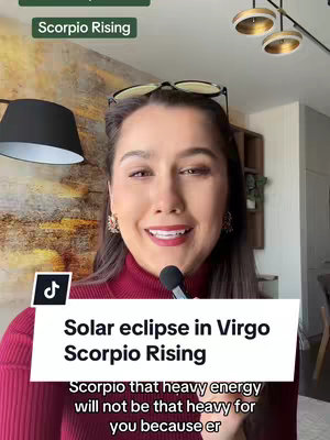 A woman with dark hair is centered in the frame, wearing a burgundy turtleneck. She's smiling and speaking into a small microphone, with a background showing an artistic wall pattern and a modern lighting fixtures. Text overlays at the top and bottom describe the video's astrology theme related to Scorpio and Virgo.