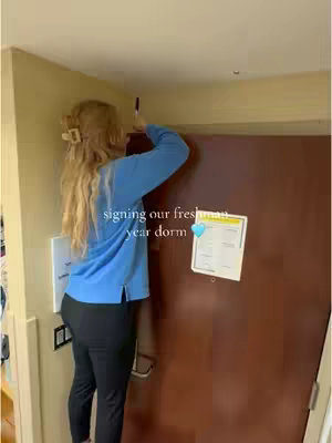 A young woman with long blonde hair is standing on her toes, writing on a door. She is wearing a light blue long-sleeved shirt and black pants. The door is brown, and there is a white sign and a door stop visible. The background is a light tan wall.
