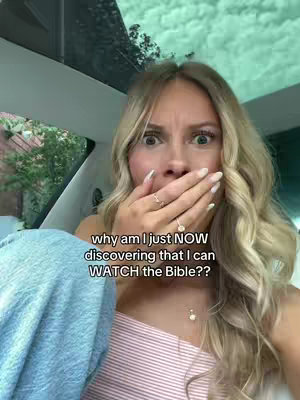 A young woman with blonde hair and a surprised expression, covering her mouth with one hand while sitting in a car. The background suggests a sunny outdoor setting, visible through the windshield and windows.
