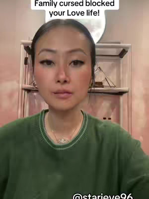 A woman with a close-up shot facing the camera. She is wearing a green sweater, has long eyelashes, and a necklace. The background is a room with a shelf containing various objects and a blurred moon shape.