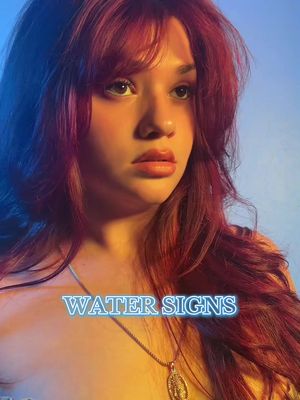 A medium shot of a young woman with long red hair, looking slightly off-camera with a contemplative expression. She wears a silver necklace, and the text 'WATER SIGNS' is overlaid on the image. The background shifts from shades of blue to a warmer yellow. The lighting is soft and diffused.