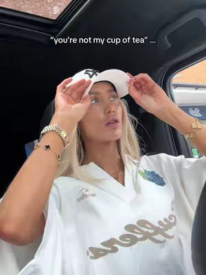 A woman is seated inside a car, looking upwards with a surprised expression, wearing a baseball cap, a white top, and jewelry. The sun roof is open, and text overlays the image.