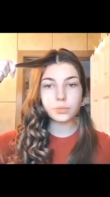 A young woman with long brown hair, styled with one side in loose curls and the other side straight. She is wearing a red shirt and holding what appears to be a flat iron to the straight side of her hair. The background is a neutral room interior.