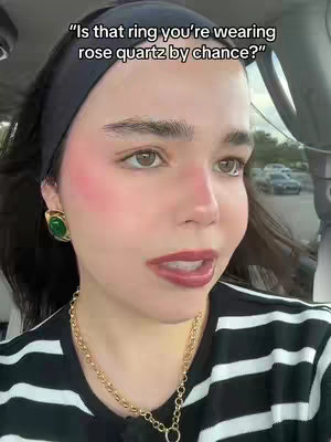 The cover image features a close-up shot of a woman inside a vehicle, likely a car. She has dark hair pulled back with a black headband and is wearing large green and gold earrings, alongside a gold necklace. Her face is framed by dramatic blush, and she has full, red lips. Text overlay reads, "Is that ring you're wearing rose quartz by chance?"