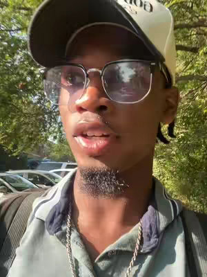 A selfie-style video cover image featuring a close-up of a light-skinned Black man wearing a black cap and glasses. The man is looking slightly off-camera and speaking, with a glimpse of green foliage and parked cars visible in the background. He has a goatee and a silver chain, with a pale complexion. The background is blurred, directing focus to the man's face. The lighting seems natural, possibly outdoors, with a warm tone.