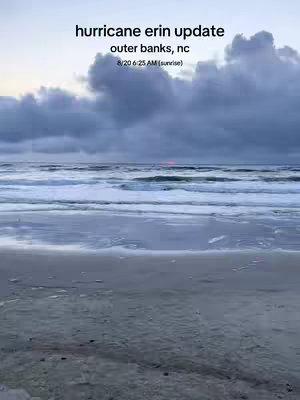 The image shows a beach scene at dawn. The foreground is the wet sand, leading to the water. The waves are visible, and the sky is filled with clouds, with the sunrise peeking through the clouds in a reddish hue. Text overlayed on top of the image states "hurricane erin update outer banks, nc 8/20 6:25 AM(sunrise)".