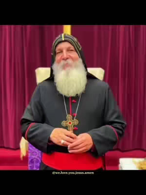 A medium shot features an older man seated in a chair, facing the camera. He has a white beard and wears a black robe with red trim and a hood. A large cross pendant hangs around his neck. The background consists of burgundy curtains.