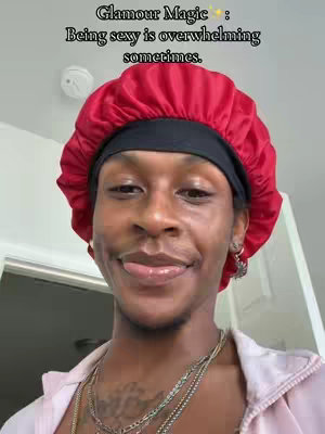 A person with dark skin is smiling with their tongue out and wearing a red satin cap. They are also wearing several necklaces. The background suggests an indoor setting. Text overlaid at the top says "Glamour Magic Being sexy is overwhelming sometimes."
