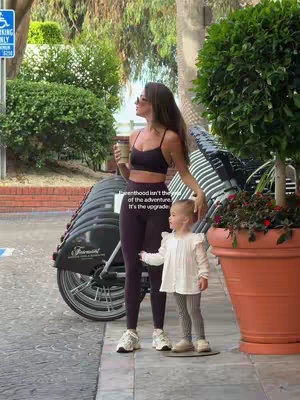 A woman and a young child stand on a stone path in front of a row of parked bicycles. The woman holds a coffee cup and they both look out of frame. The background includes greenery and a glimpse of street signs.