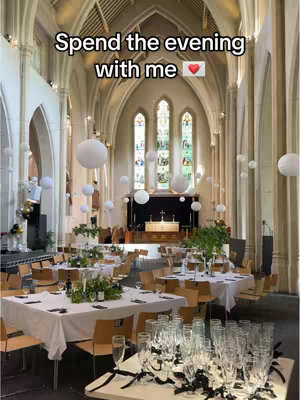 An interior shot of a large church space set up for a social gathering. Tables with white tablecloths and elegant centerpieces are arranged throughout the room, and tall arched windows with stained glass provide a backdrop. White paper lanterns hang from the ceiling, and the overall atmosphere suggests a celebratory event.