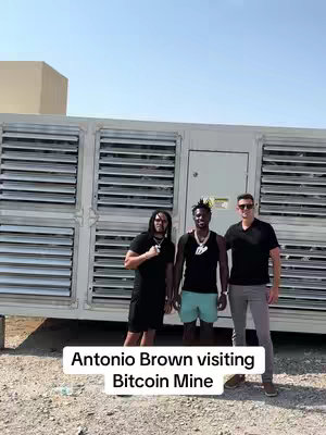Three men stand outside with a large industrial-looking structure behind them against a clear, sunny sky. One man is wearing a black t-shirt. Another man is wearing a black tank top with turquoise shorts and the third man is wearing a black t-shirt and sunglasses. The text overlay "Antonio Brown visiting Bitcoin Mine" is against a light grey background.