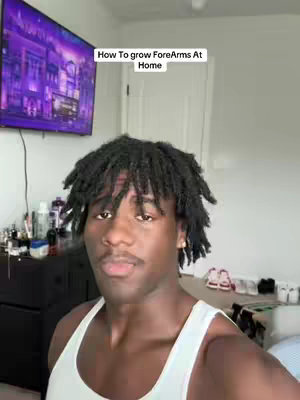 A man with dreadlocks is centered, looking into the camera, above him is text that reads "How To grow ForeArms At Home" The man is wearing a white tank top. Behind him is a room with a large tv visible, a door to the right and a dark dresser with various items on it.