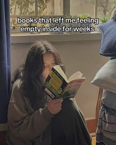 A young woman sits on the floor near a window, engrossed in a book. The lighting is soft, suggesting a peaceful indoor setting. Additional elements includes pillows, a window and a hint of greenery adds to the cozy indoor scene.