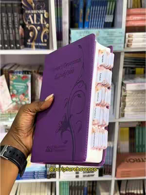 A purple Bible is presented, held in a hand against a backdrop of bookshelves filled with books. The Bible is open and shows the tabs of the pages. The hand holding the Bible is dark-skinned, and the background features various other books. The overall setting suggests a bookstore or a personal collection.