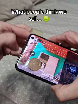 The image shows a person holding a phone displaying an app screen. The phone is held against a somewhat blurred background, possibly a bed or soft surface. Overlaid text reads, 'What people think we sell...'. The app screen seems to show elements of a game interface or perhaps another application.