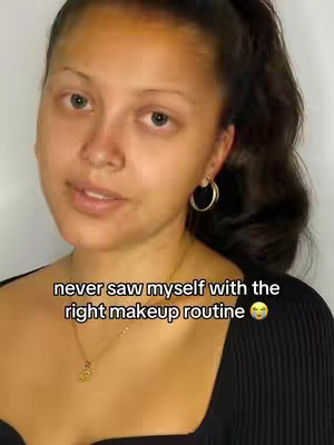 A young woman with light brown skin is centered in a vertical shot. She has dark, wavy hair pulled back into a ponytail. Gold hoop earrings are visible. She is wearing a black top and a gold necklace with a charm. Text overlayed on the image reads, "never saw myself with the right makeup routine ", along with a rolling-eyes emoji. The background is blurred, and the lighting appears natural.