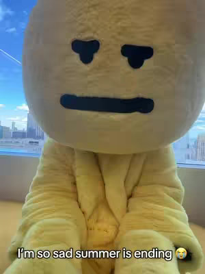 The image shows a person in a large, yellow costume, resembling a smiling, round-faced character. The costume has small black eye details on the face and a large mouth. The background shows tall buildings. The text overlay says, "I'm so sad summer is ending."