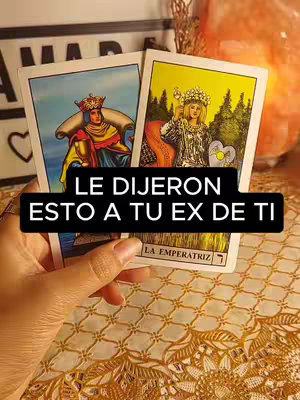 A hand holding two tarot cards, featuring illustrations of figures on a yellowy-cream background. The cards are placed against a background of patterned cloth and a blurred setting. In the background, there's a lamp or decorative item casting a soft glow and a sign.