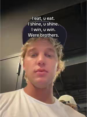 A selfie-style shot featuring a young man with blonde hair, wearing a white t-shirt and a dark blue baseball cap. The image is lit with indoor lighting, and the man is directly facing the camera. Text overlayed on the image reads, "I eat, u eat. I shine, u shine. I win, u win. Were brothers."