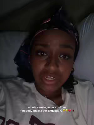 A medium close-up shot of a Black woman lying in bed, speaking directly to the camera. She has dark curly hair with a patterned headscarf, and is wearing a white t-shirt. Text overlayed at the bottom asks, 'who is carrying on our culture if nobody speaks the language??' with heart emojis.