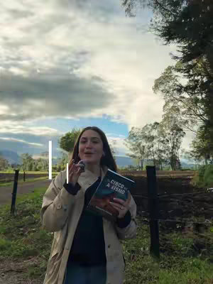 A woman stands outdoors, holding a book and a microphone. The background includes a rural scene with trees and a cloudy sky.