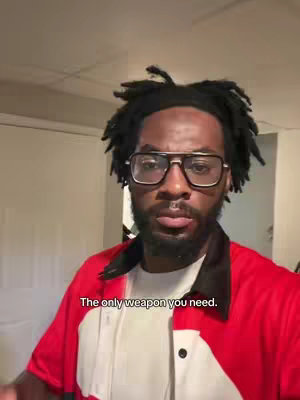 A man with dark skin and thick dreadlocks is centered in a close-up shot. He wears large, black-rimmed glasses and a red shirt with a white undershirt. Text overlayed at the bottom of the image reads, 'The only weapon you need.' The background is blurred, showing what appears to be a white wall or door.