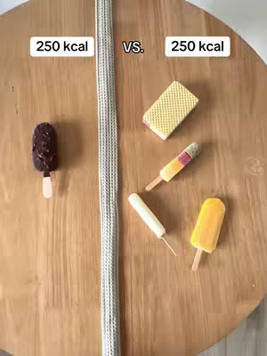 A top-down shot of a light wooden surface, likely a table or countertop. A wide, pale grey, textured object (possibly a placemat or segmented rope-like item) bisects the image vertically. On the left, a small ice cream bar on a stick. On the right, five different ice pops and bars are arranged.  Above the treats, the text "250 kcal" is displayed on both sides of the vertical divider, and “VS.” is in the middle.