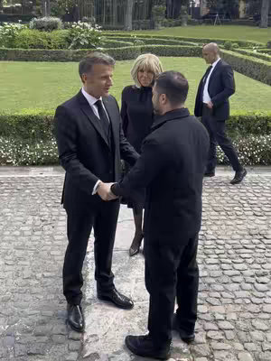 The cover image shows three men and a woman, presumably political figures, in a park setting. Two men in suits are shaking hands in the foreground, with another man and a woman standing nearby. The background features trimmed hedges and foliage.