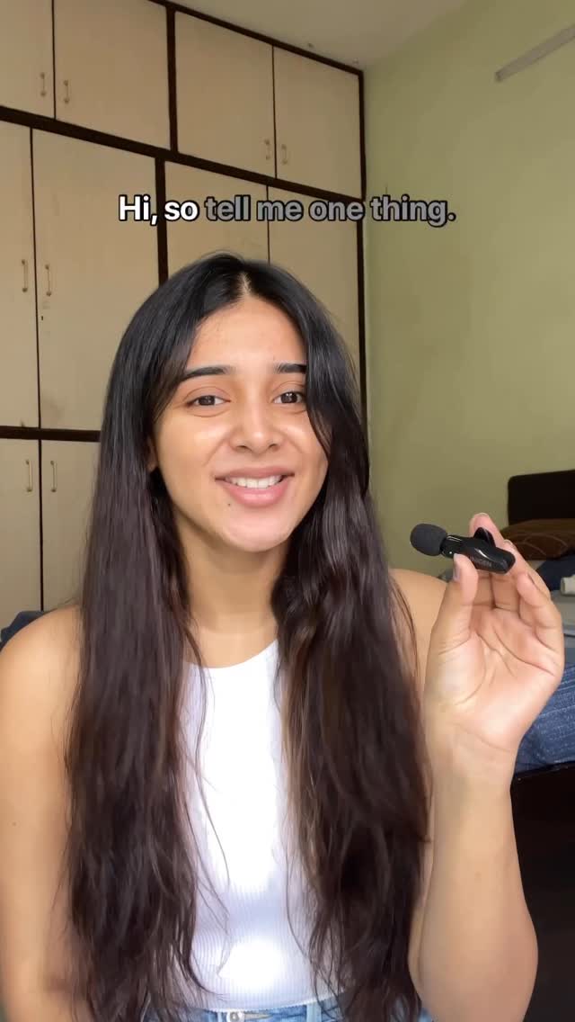 A woman with long, dark hair smiles at the camera while holding a small microphone. Behind her, a neutral-toned room with white cabinets is visible. The hook text, "Hi, so tell me one thing" arcs above her head.