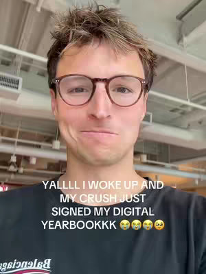 A selfie-style shot of a young man with brown hair and glasses, smiling slightly. Text overlayed on the image reads, 'YALLL I WOKE UP AND MY CRUSH JUST SIGNED MY DIGITAL YEARBOOKKK'. The background is slightly blurred, showing an indoor space with white ceiling elements.
