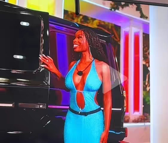 A young woman in a light blue dress stands next to a black car, smiling and looking to her right. She has dark hair and is wearing a necklace. The setting appears to be outdoors with some blurred background elements visible.