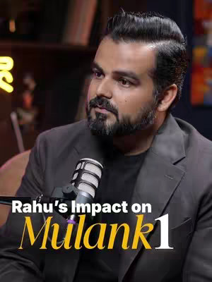 A man with dark hair and a beard is seated in a studio setting. He is wearing a dark blazer over a black shirt and is positioned next to a microphone. Colored ambient lights illuminate the background, which includes partial views of bookshelves. The text overlay at the bottom of the image reads, 'Rahu's Impact on Mulank 1'.