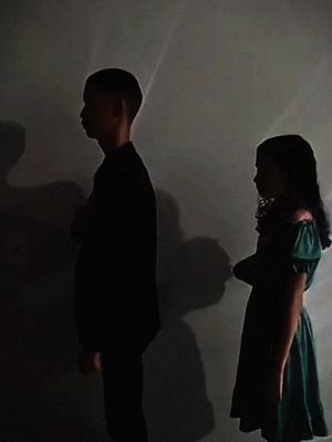 A grayscale photo shows two children standing against a plain wall. One appears to be a slightly blurred shadow with only his profile visible. The other is a girl, also in profile, wearing a dark dress. The lighting suggests a spotlight on their silhouettes.