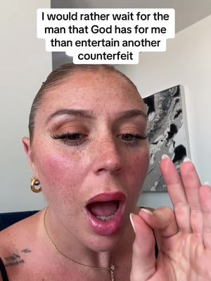 A selfie-style shot featuring a woman with fair skin and prominent freckles, set against a blurred indoor backdrop. The woman is looking directly at the camera with her mouth open wide, conveying surprise or emphasis. She is wearing gold hoop earrings and a delicate necklace. Added text floats above her head while she is making a hand gesture.