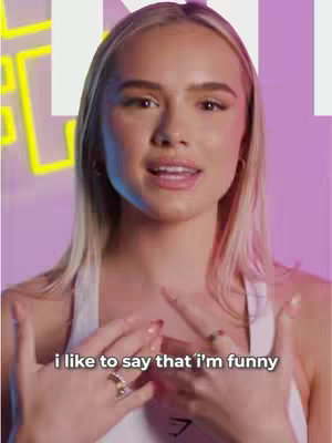 A woman with blonde hair and a white top, smiling. Text overlayed reads, 'i like to say that i'm funny.' The background is blurred, with a pink and purple gradient and a neon 'K' logo.