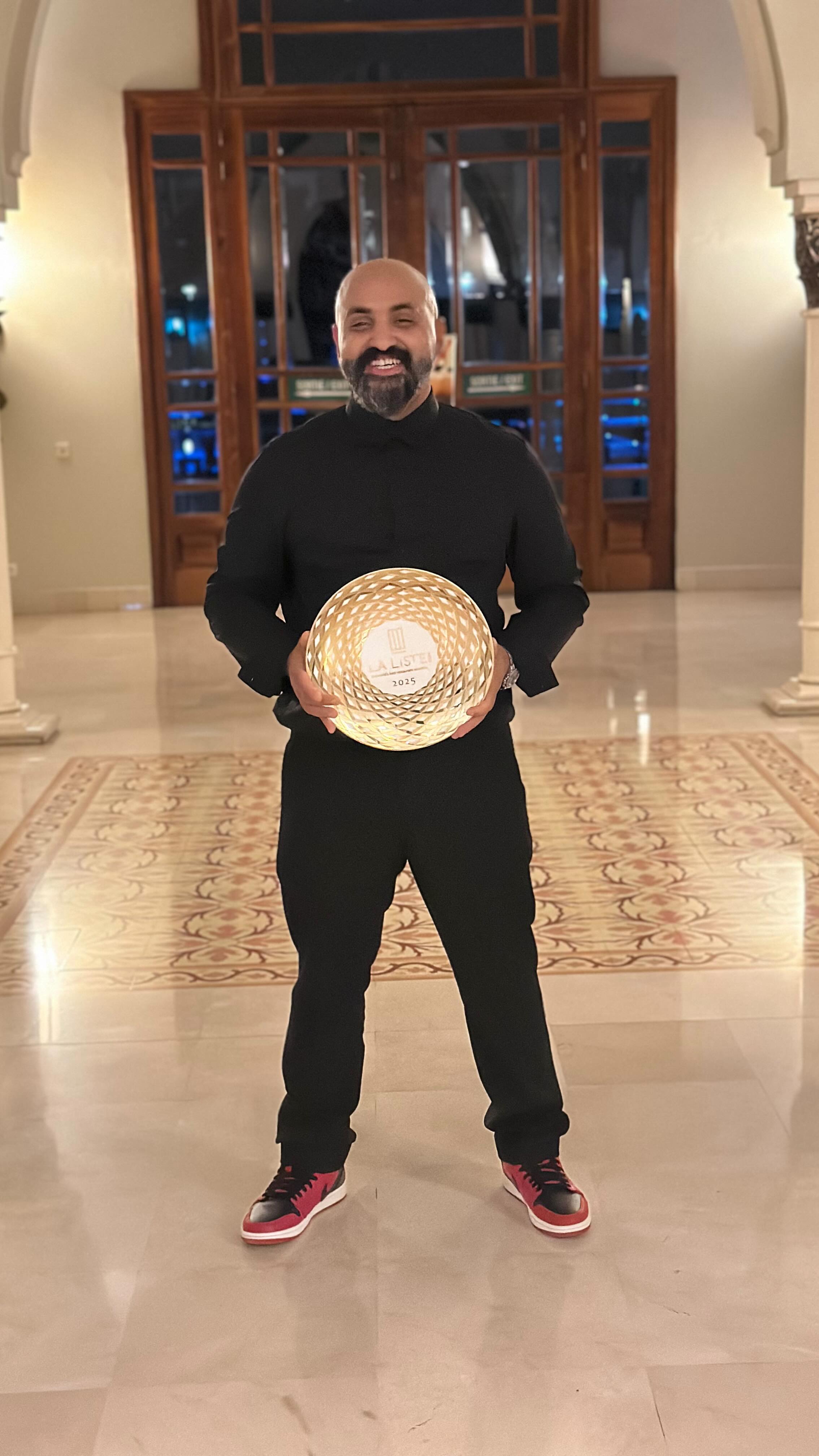 A full-body shot of a smiling man holding up a gold award. He is in a well-lit, ornate hallway with a patterned floor. The man is wearing a black shirt and pants, with red-and-black sneakers. The award is circular with "LA LISTE 2025" printed on it.