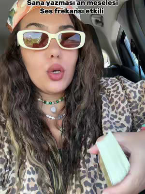 A selfie-style shot featuring a woman in a car. She wears large, rectangular cream-colored sunglasses and a patterned scarf and shirt, with long, wavy brown hair. Text overlay, appears at the top. She is looking at the camera.