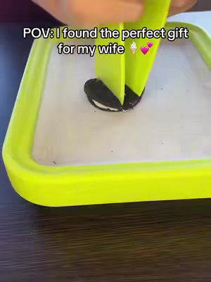 The image shows a bright green square tray with a white interior, black cookie with white cream in the middle and two green paddles are holding the cookie and scooping the cookie cream.